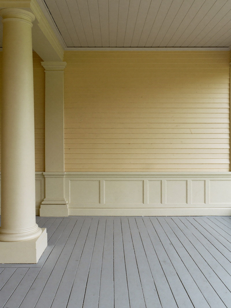 A restored front porch on an established Natick colonial: square painted columns in soft cream, warm gray painted fir floor, cream beadboard ceiling, pale
