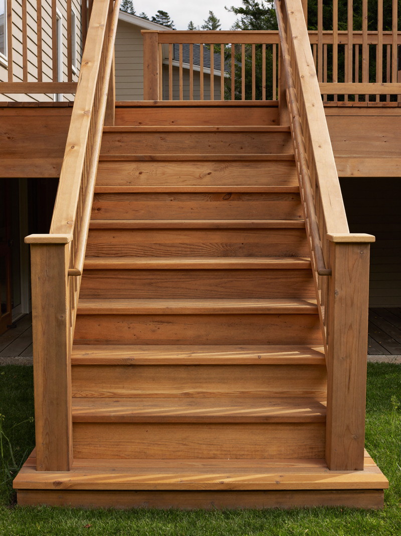 Rebuilt wooden deck stairs on an established suburban colonial: warm cedar-toned treads, natural wood risers and balusters, smooth oiled handrail, green
