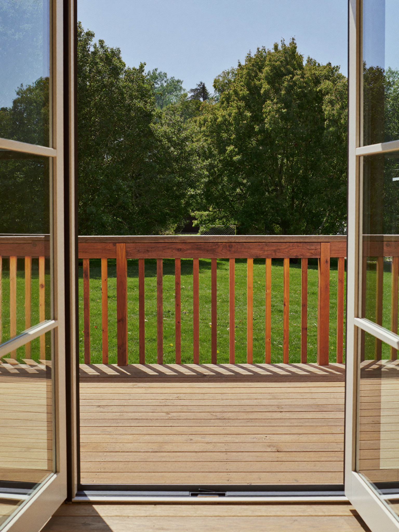 Tall photograph through open french doors onto a rebuilt suburban deck: warm cedar-toned boards, natural wood railing with smooth oiled top rail, green