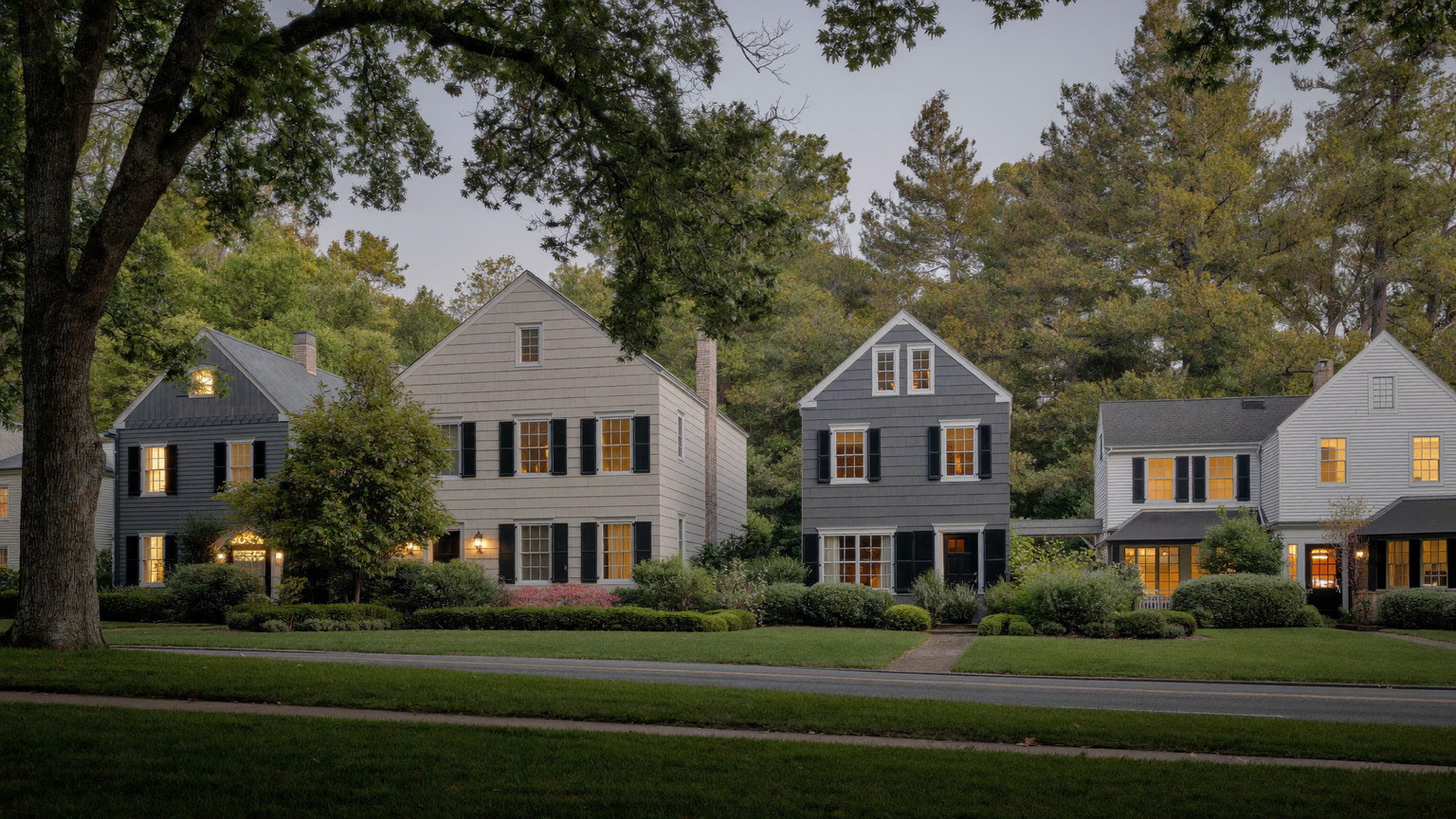 A quiet leafy Natick Massachusetts residential street in early evening: established detached colonials and capes set apart on deep green lawns, dark
