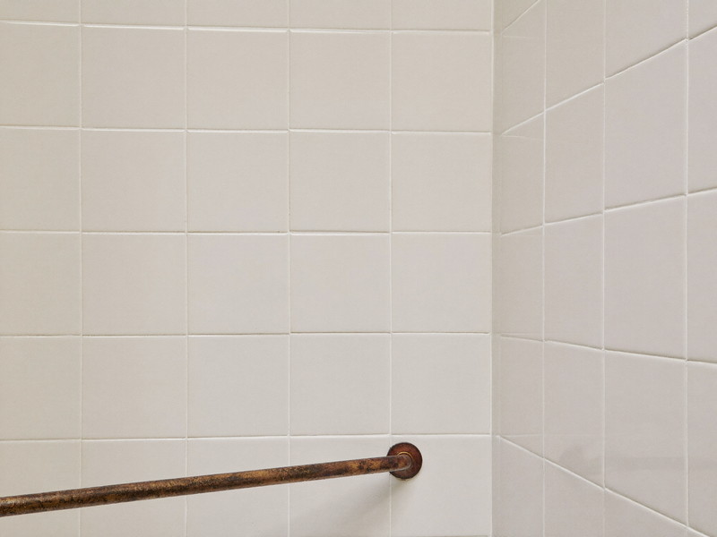 Freshly regrouted warm white glazed ceramic tile in a suburban shower corner: clean bright grout lines, every tile flat and straight, aged bronze fitting