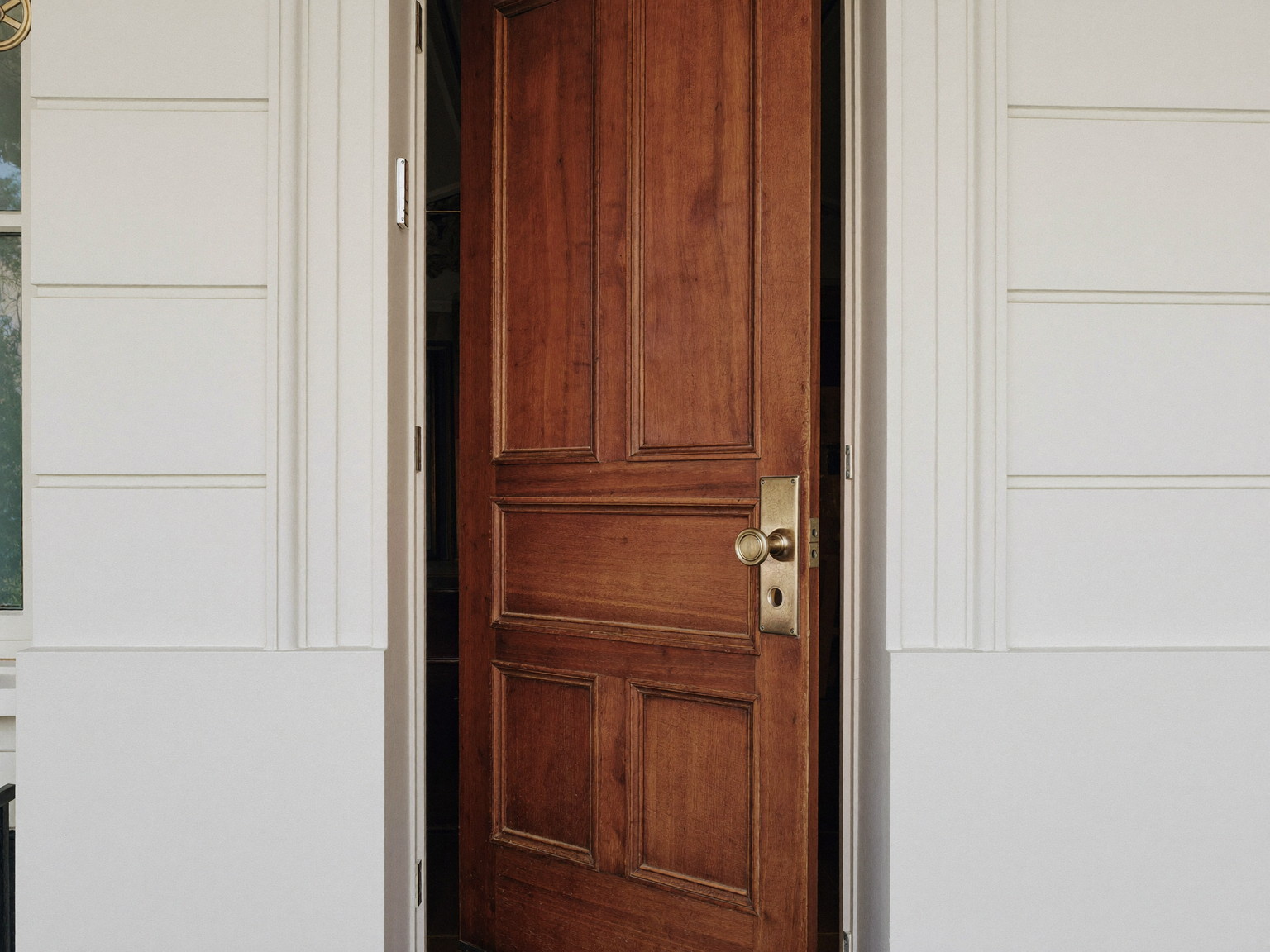 A traditional stained wooden front door standing slightly open in its freshly serviced frame inside a colonial entry: raised panels visible, smooth