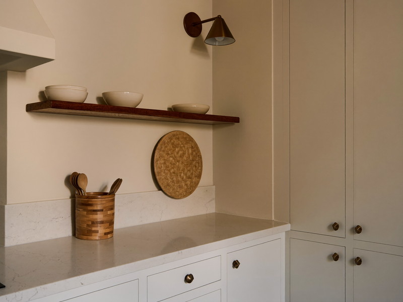 A finished family kitchen corner at evening: warm white inset cabinets with aged bronze knobs, pale quartzite counter completely bare, cherry wood shelf