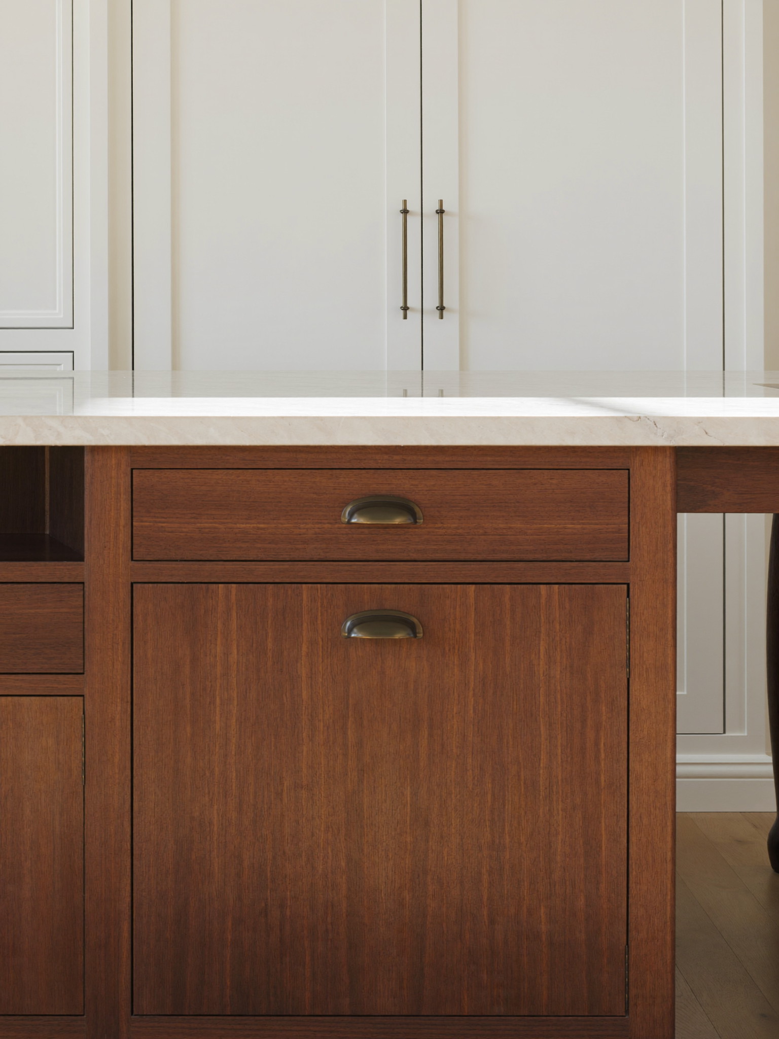 A generous cherry wood kitchen island with a pale quartzite top and simple turned legs, aged bronze hardware with smooth faces, warm white inset cabinets