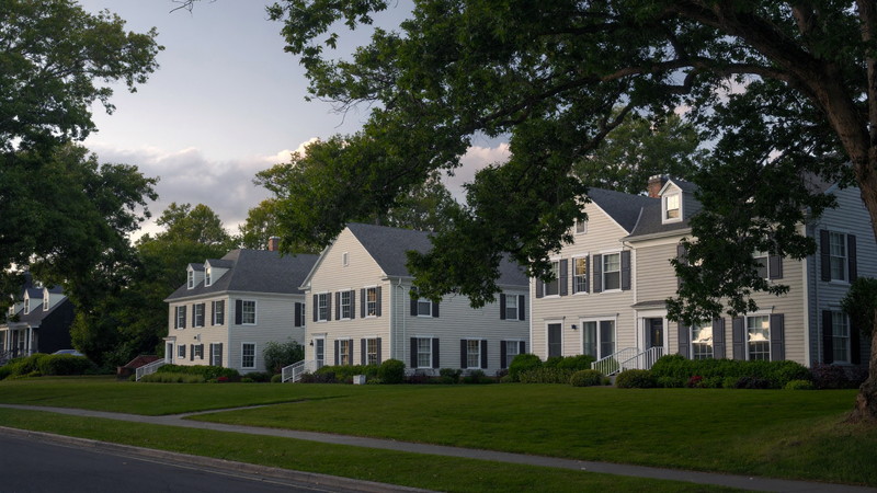 A leafy Natick Massachusetts residential street in soft evening light: established detached colonials and capes set apart on deep green lawns, dark