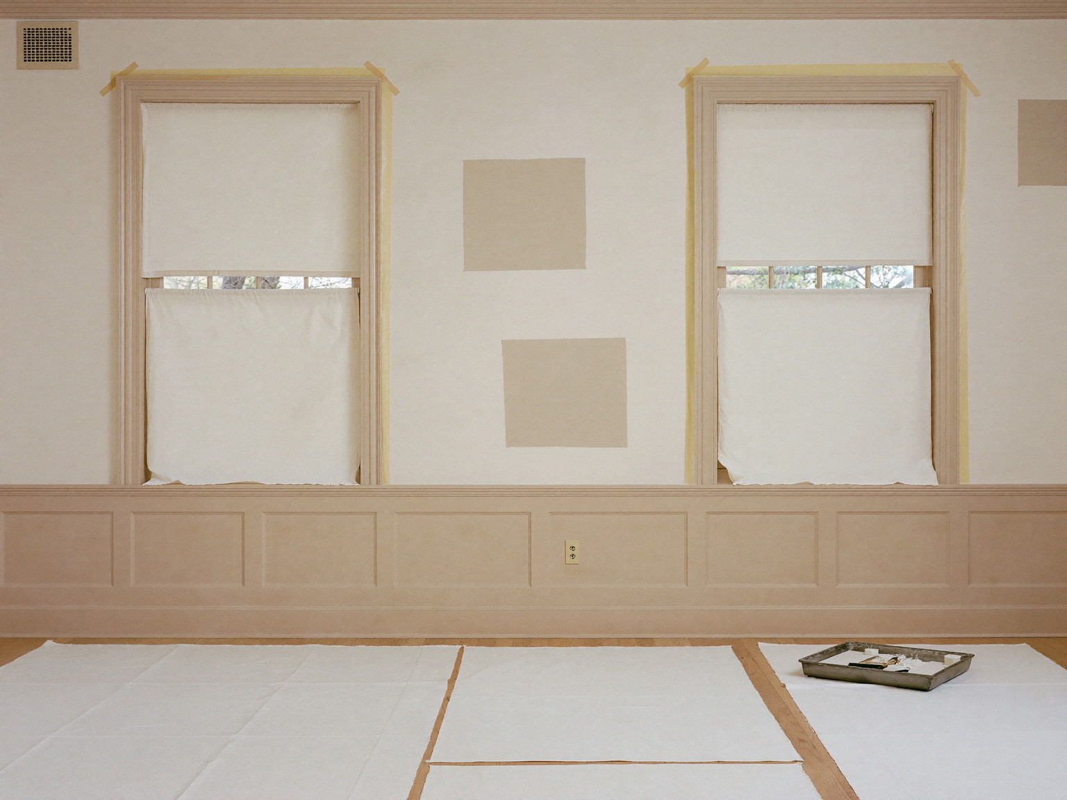 Interior painting prep, seen from across an established colonial room: clean canvas drop cloths spread over the oak floor, window casings masked with