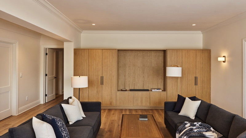 A warmly finished lower-level family room beneath an established Needham Massachusetts colonial: warm ivory walls, white oak built-in media wall with