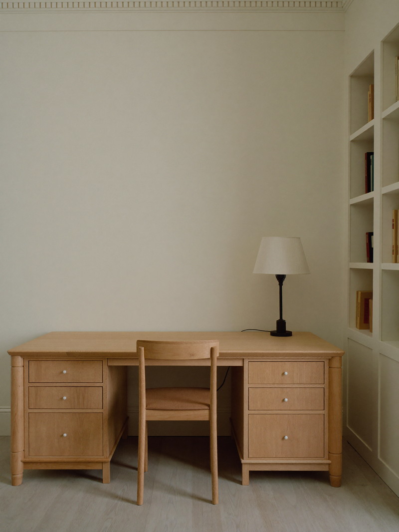 A finished lower-level home office in a renovated colonial basement: built-in white oak desk with plain drawer faces against a warm ivory wall, simple