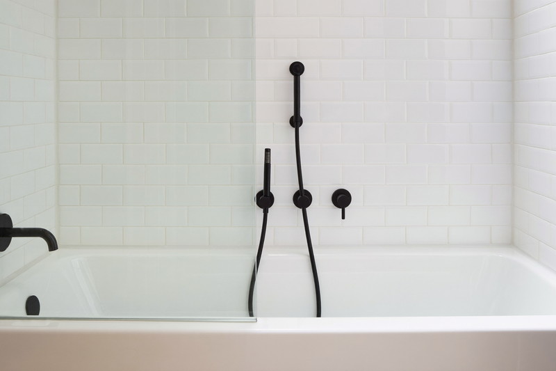 A refined tub and shower combination in a suburban family bathroom: white tub, warm white stacked tile surround, matte black fixtures, clear glass screen,