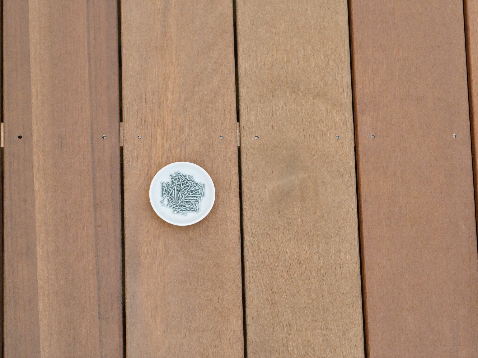 Overhead macro of new composite decking boards stacked neatly, smooth bare surfaces, beside them a small plain white ceramic dish holding straight smooth