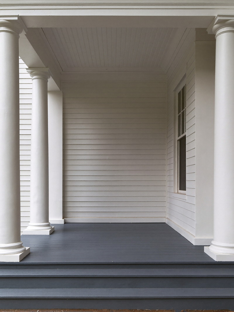 A restored front porch on an established Needham colonial: square white columns, warm gray painted fir floor, white beadboard ceiling, white clapboard