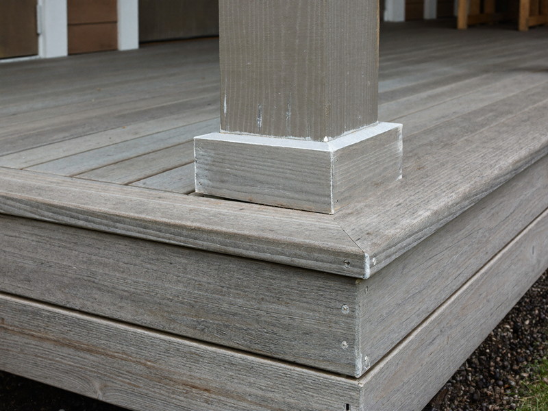 Close-up of an aged deck post base on an established suburban home before repair: layers of weathered gray finish, soft wood grain at the bottom, honest