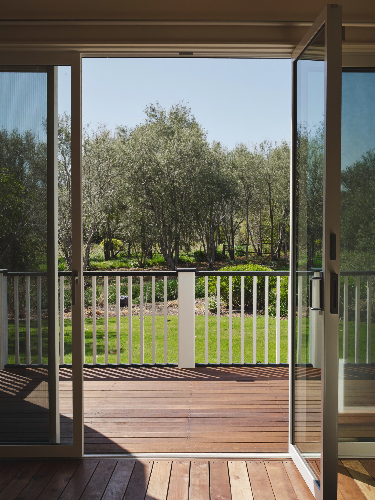 Tall photograph through open sliding doors onto a newly built suburban deck: warm brown composite boards, white balusters with black top rail, green lawn