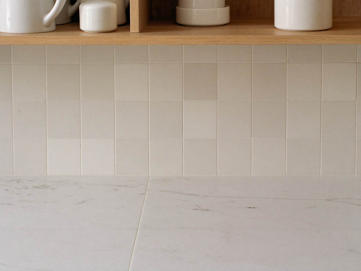 Macro photograph of a honed white stone counter with faint warm veining meeting a warm white square tile backsplash, a slim white oak shelf above holding