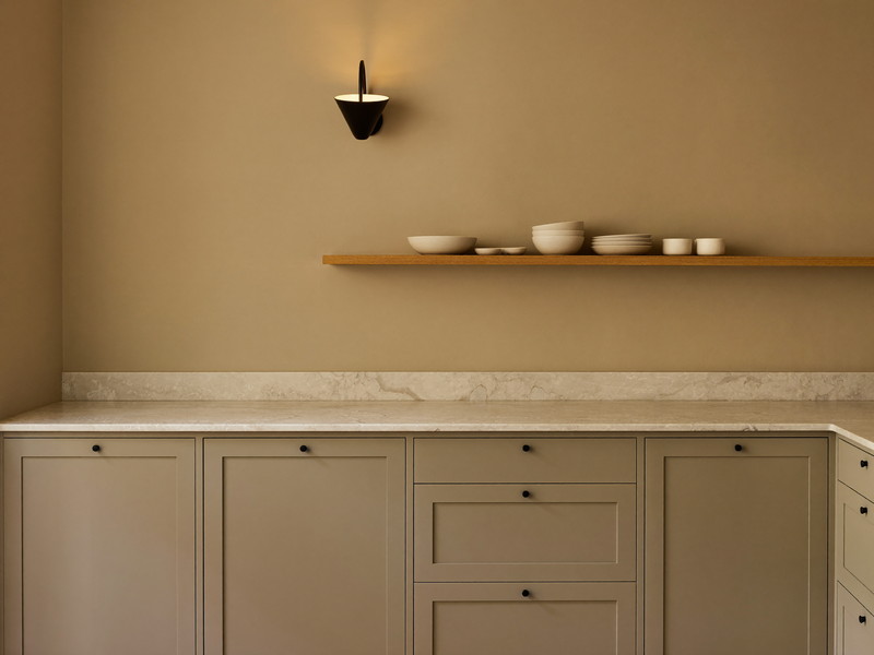 A finished family kitchen corner at evening: warm greige inset cabinets, honed white stone counter, white oak shelf lit warm by a small black sconce, calm