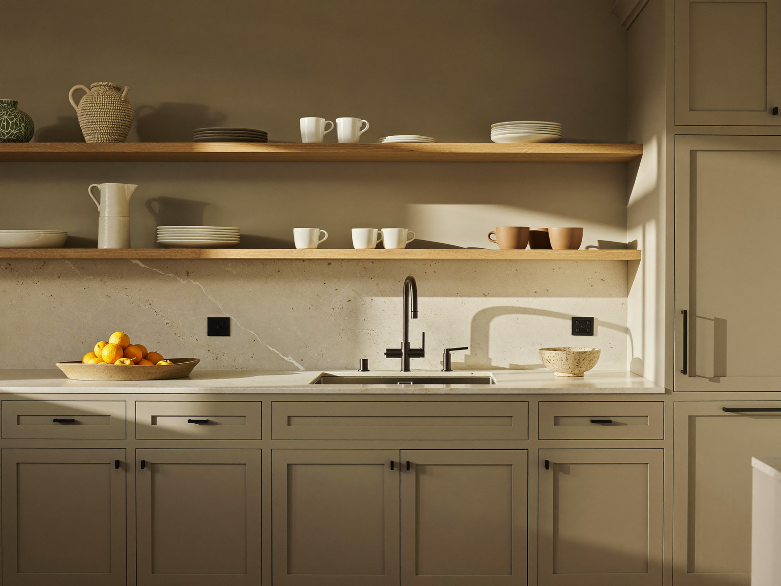 A completed suburban family kitchen seen straight on: warm greige inset cabinetry, honed white stone counters, white oak open shelf holding plain white