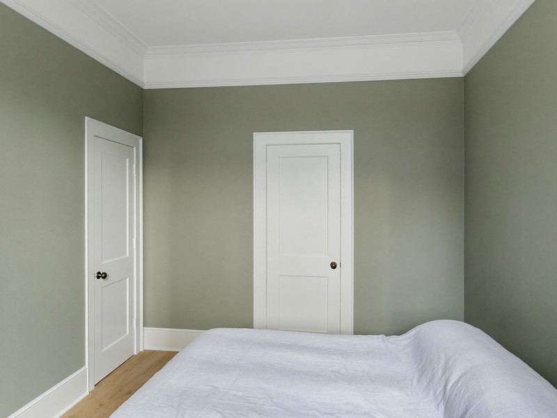 A repainted bedroom in an established Needham colonial: soft sage-gray walls, bright white trim and paneled door, smooth restored plaster in raking light,