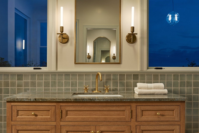 Finished bathroom at dusk with lit brass sconces, arched mirror, and sage tile glowing warm