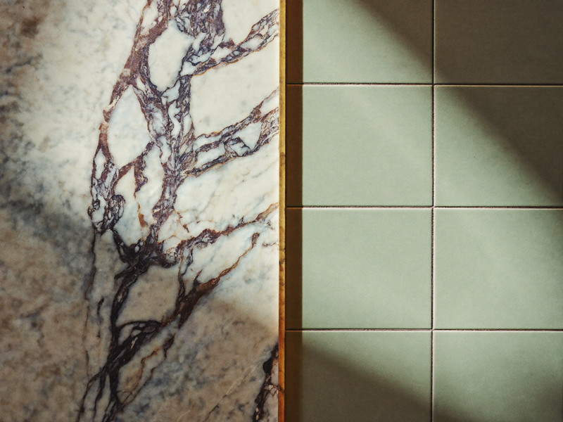 Honed carrara marble meeting sage ceramic tile with a crisp caulk line and brass trim strip