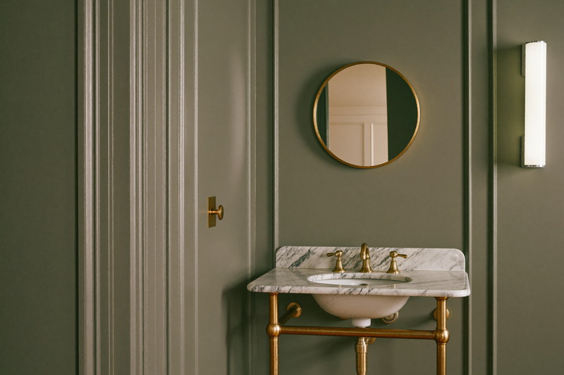 Powder room corner with a marble-topped brass washstand, round mirror, and deep sage paneled walls