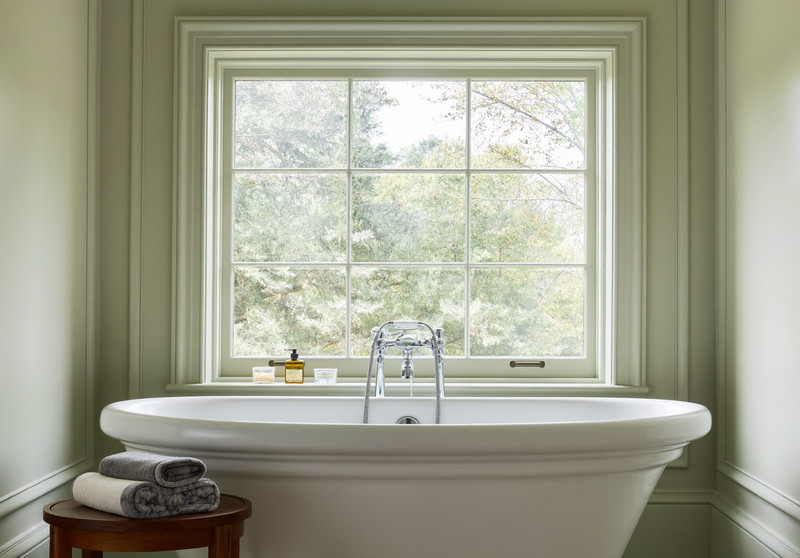 Freestanding tub in a windowed alcove with crisp white trim, walnut side table, and sage roman shade