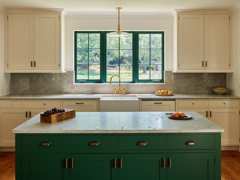 Completed New England kitchen with cream inset cabinetry, forest-green island, and a farmhouse sink under a triple window
