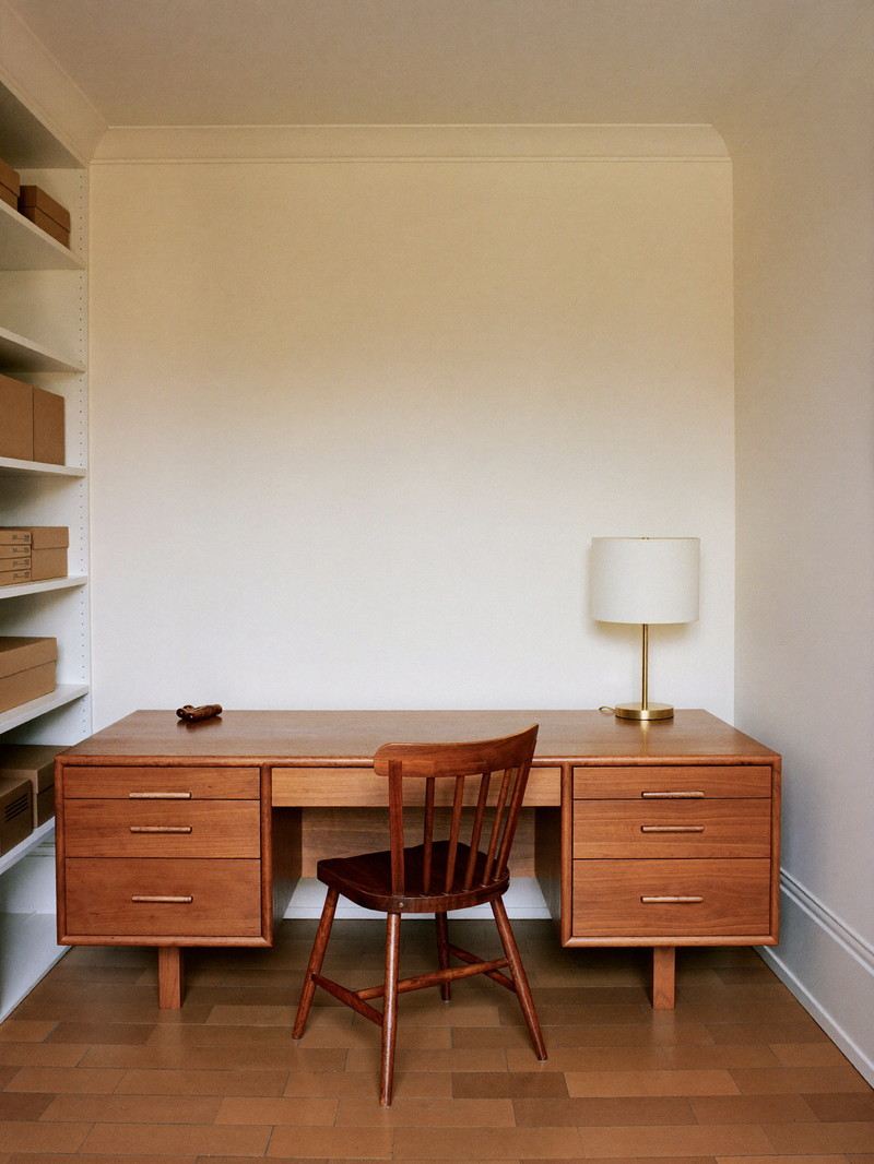 A finished lower-level home office: warm wood desk and chair against a cream wall, brass task lamp with a plain shade, neat open shelves with plain boxes