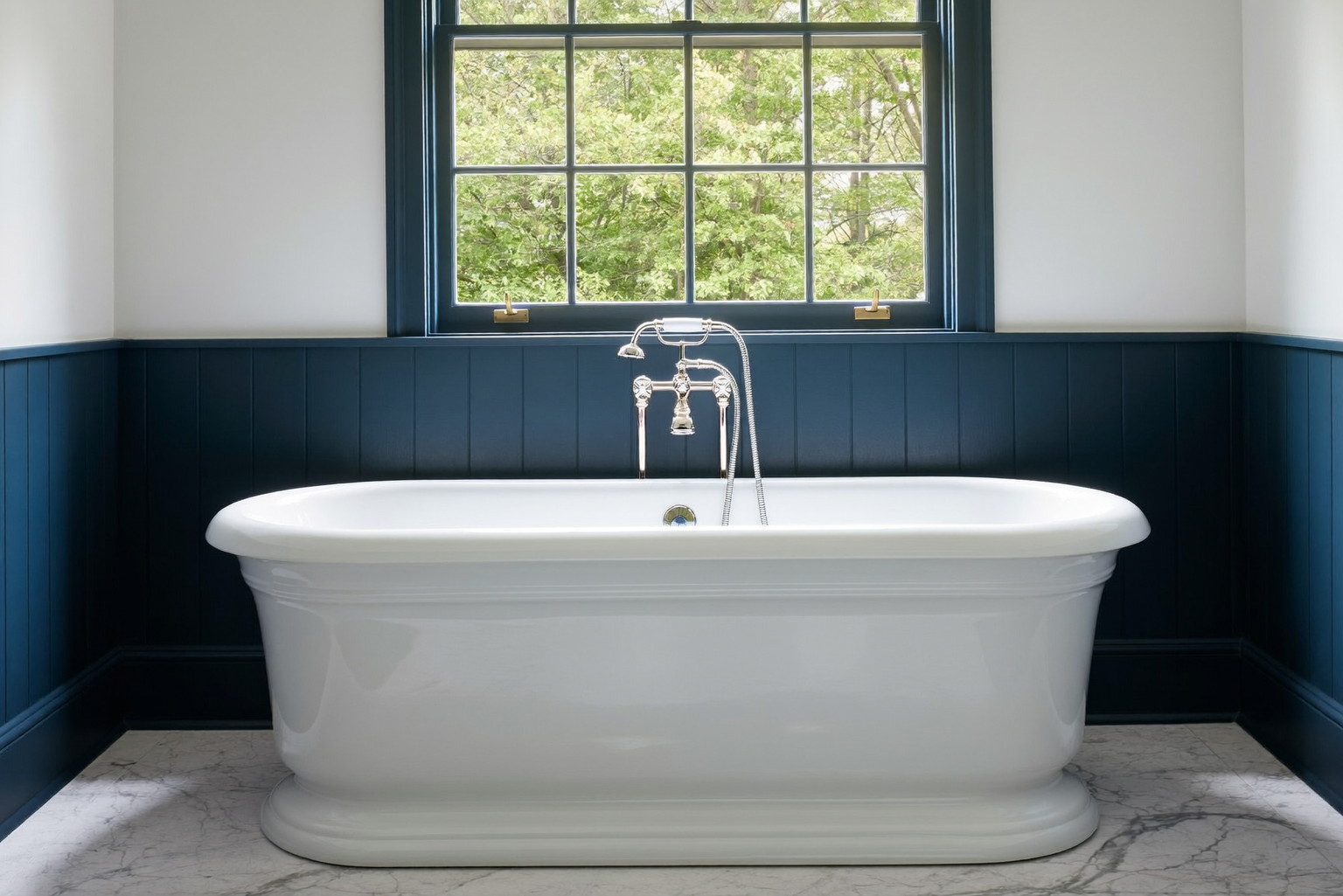 A renovated bathroom in a classic Wellesley colonial: white freestanding tub beneath a wide window with muntins, deep navy wainscot, warm white walls, polished