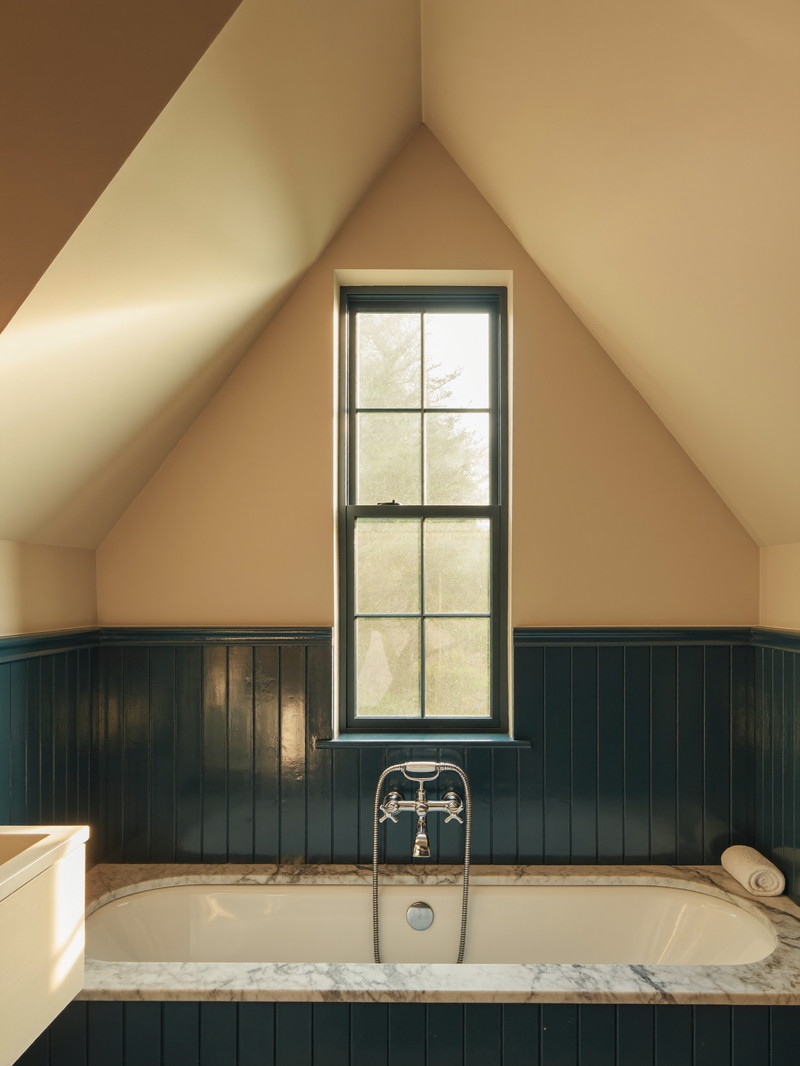 A renovated bathroom under a sloped dormer ceiling in a classic Wellesley cape: white tub, deep navy wainscot, honed marble, polished nickel, tall narrow