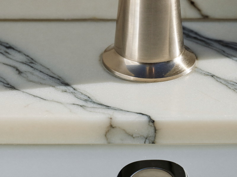 Macro photograph of honed white marble with soft gray veining meeting a polished nickel smooth-faced faucet base, shallow depth of field, bright natural light