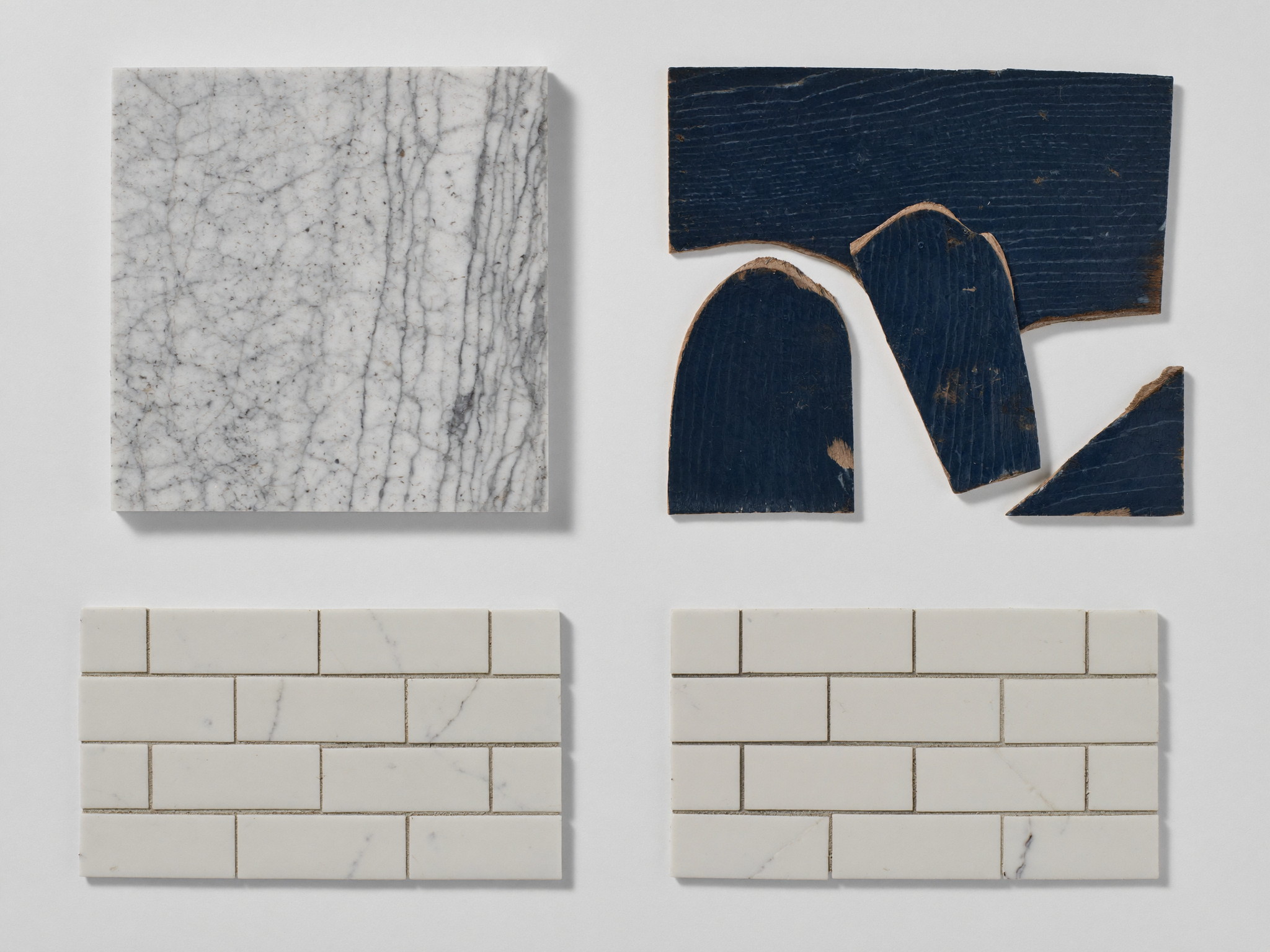 Overhead flat-lay on plain white paper: a square slab of honed white marble, sample pieces of deep navy painted wood, and white marble subway tiles, soft daylight