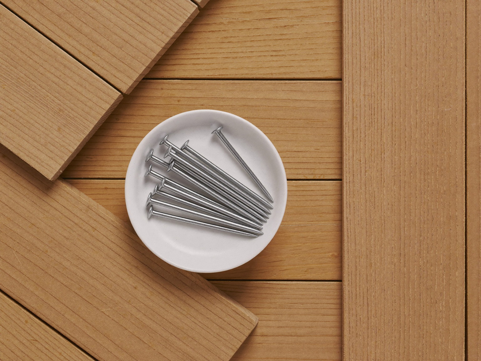 Overhead macro of new cedar decking boards stacked neatly, smooth bare wood grain, beside them a small plain white ceramic dish holding straight smooth