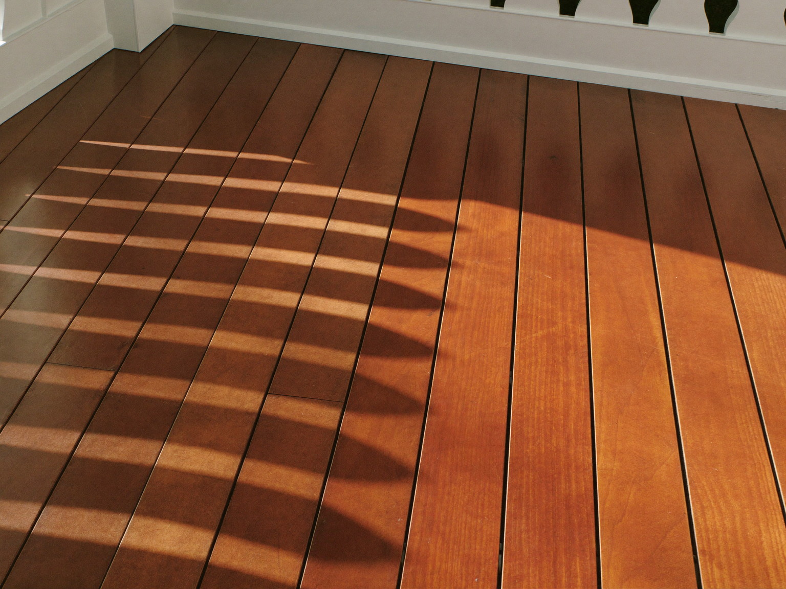Freshly sealed cedar deck boards in warm afternoon light: rich amber finish with a soft sheen on smooth grain, crisp white baluster shadows falling across the
