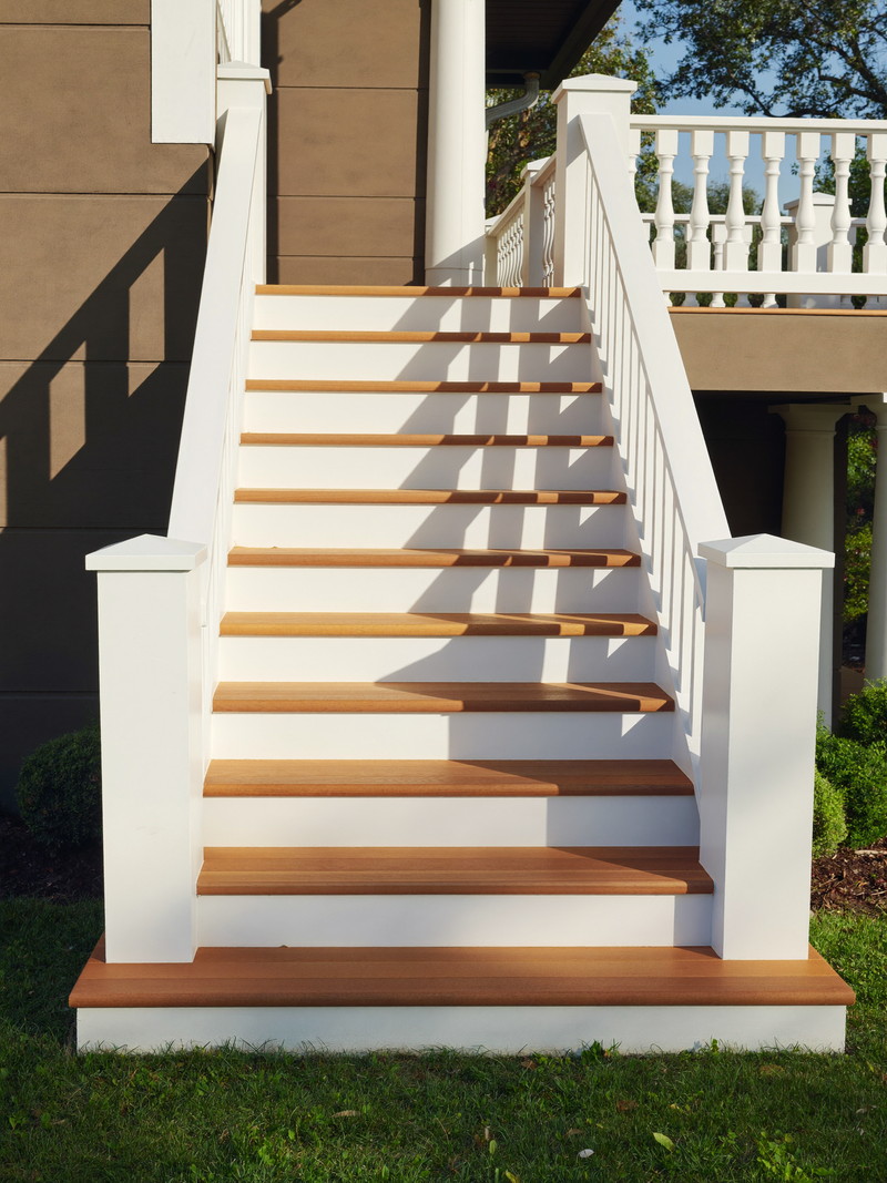 Rebuilt deck stairs on a classic suburban colonial: warm cedar treads, crisp white risers and classic white balustrade, green lawn below, afternoon light