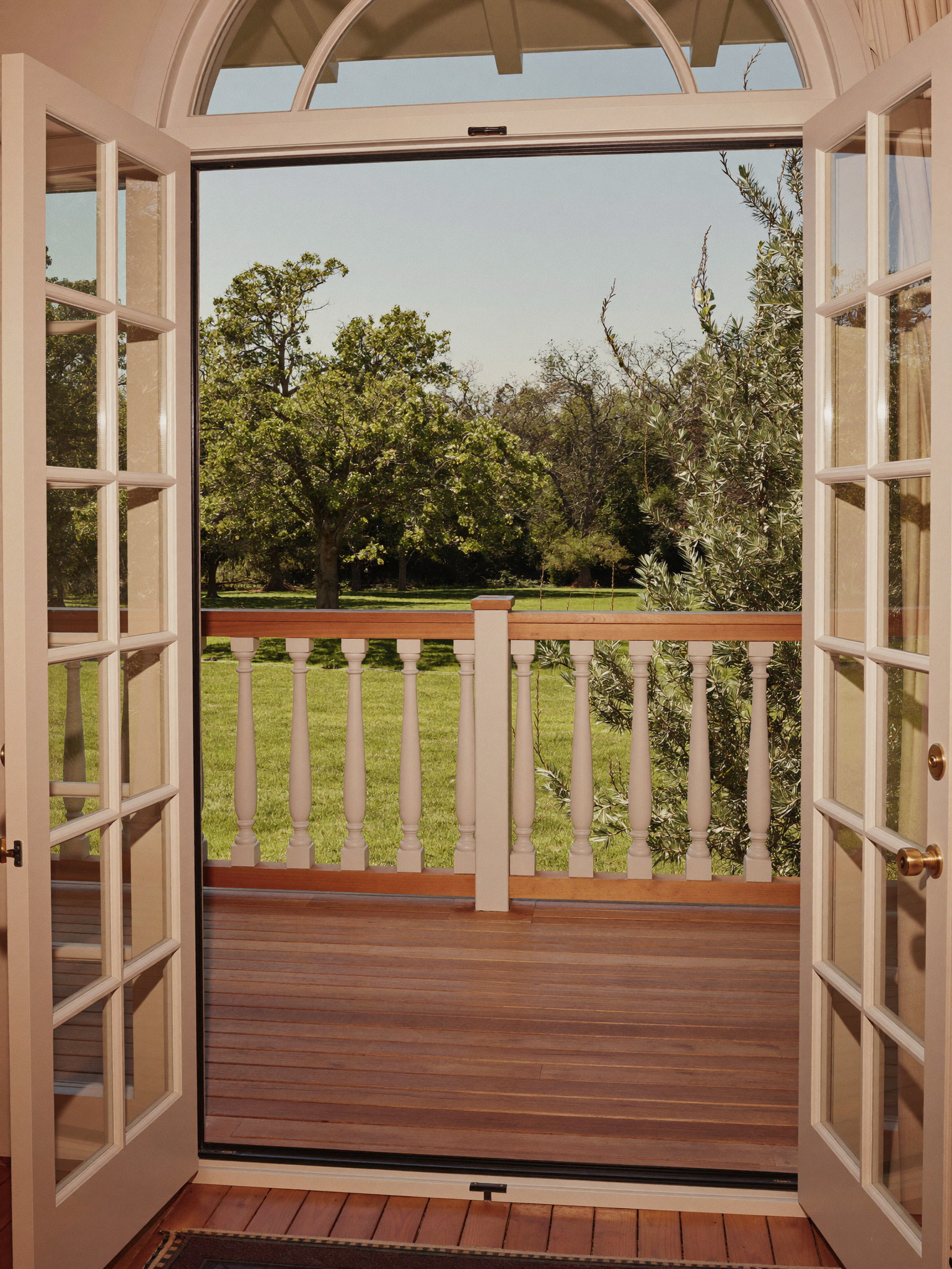 Tall photograph through open French doors with an arched transom onto a rebuilt porch: warm cedar boards, classic white balustrade, green lawn and mature trees