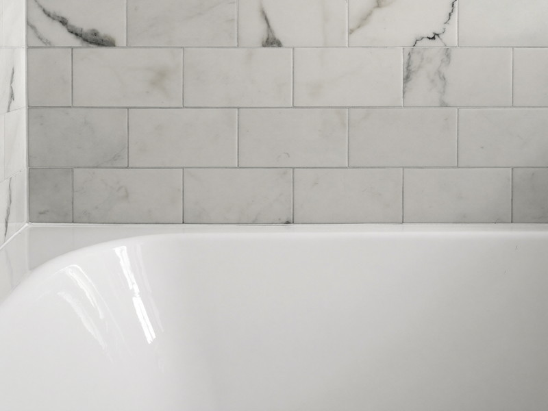 A clean bright white bathtub edge meeting white marble subway wall tile, the joint freshly finished, smooth and tidy, completely bare tub edge, soft daylight