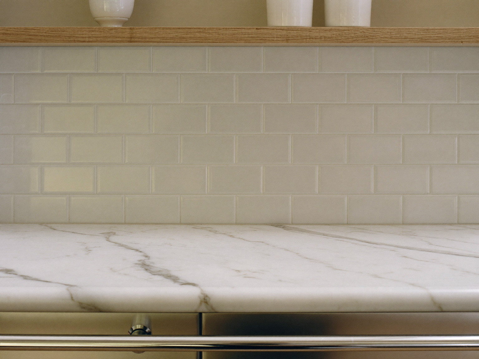 Macro photograph of a honed white marble counter with soft veining meeting a plain white beveled subway backsplash, a slim white oak shelf above holding plain