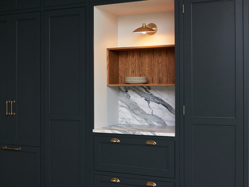 A finished classic kitchen corner at evening: deep navy inset cabinets, honed marble counter, white oak shelf lit warm by a small nickel sconce, calm and