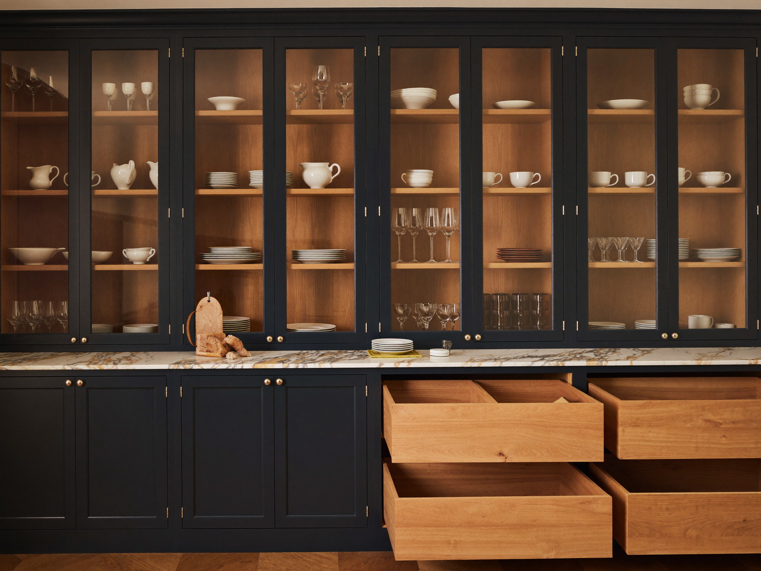 A butler pantry in a renovated colonial: floor-to-ceiling deep navy inset cabinets with glass uppers holding plain white china, honed marble counter, white oak